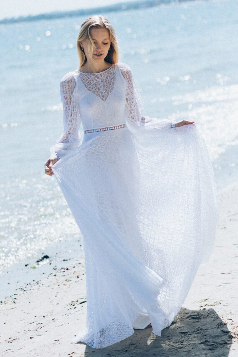 Suknia z długimi rękawami i zwiewnym trenem widoczna na plaży.