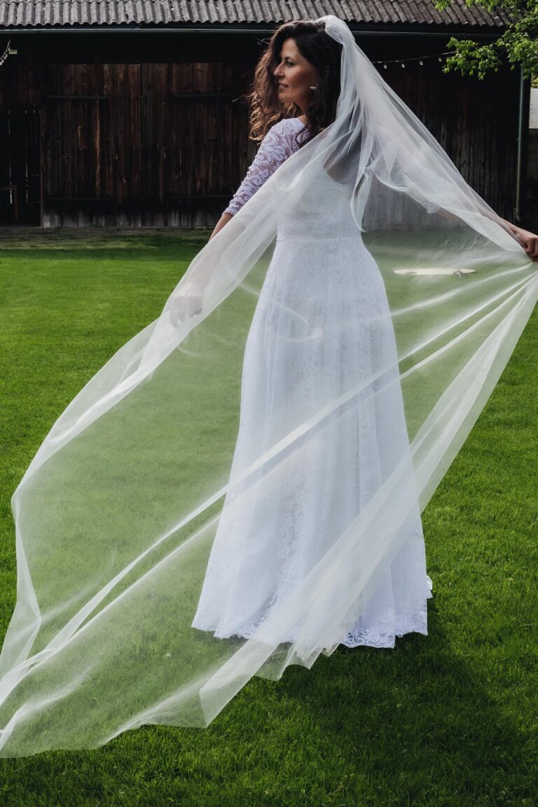 Suknia ślubna z długim trenem i welonem, widziana z tyłu na zielonej trawie.