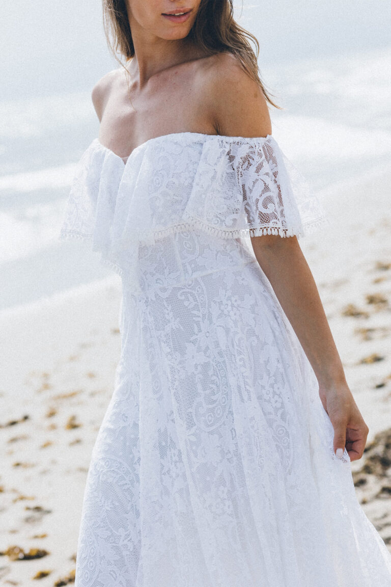 Suknia ślubna z odkrytymi ramionami i koronkowymi rękawami, wykonana z białego materiału.