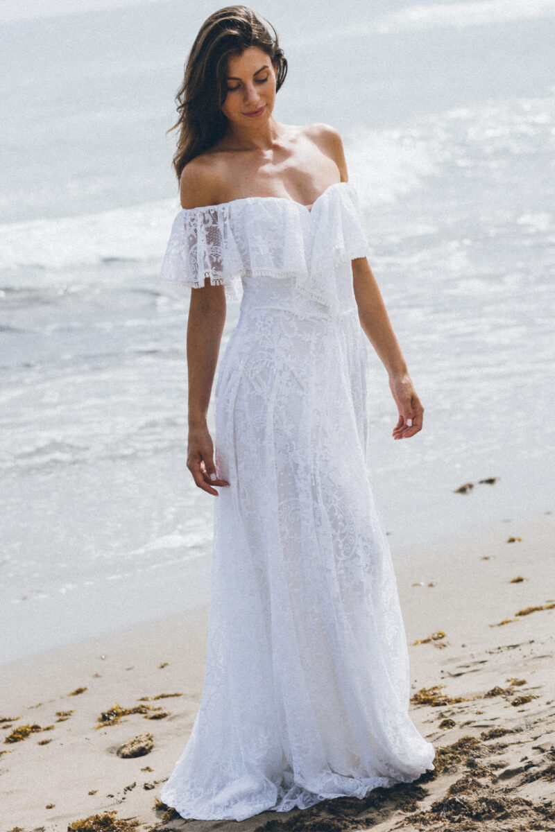 suknia ślubna z odkrytymi ramionami, wykonana z koronkowego materiału, modelka stoi na plaży